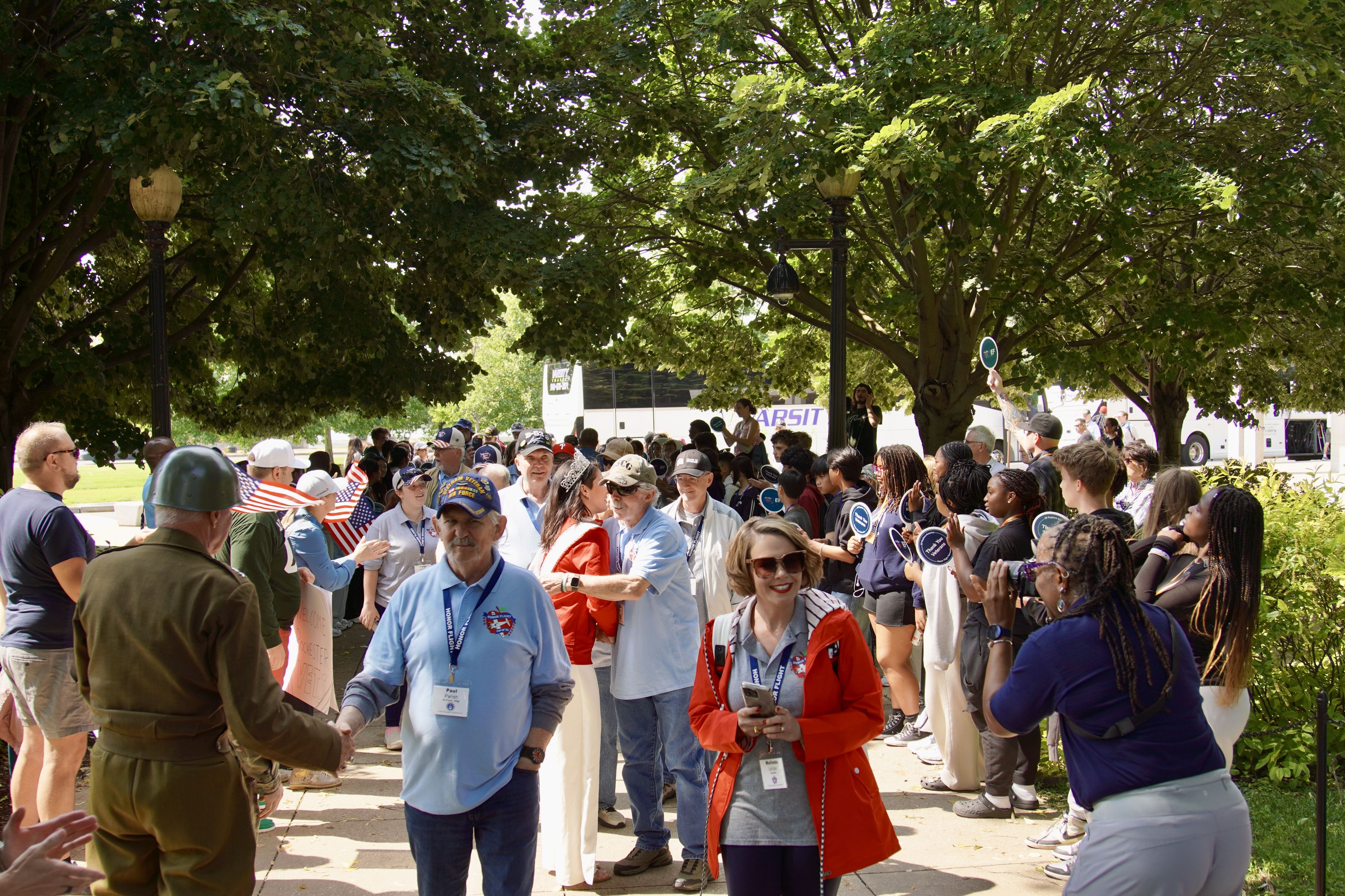 A group of people, some in uniforms, gather outdoors under trees, engaging in conversations and taking photos.