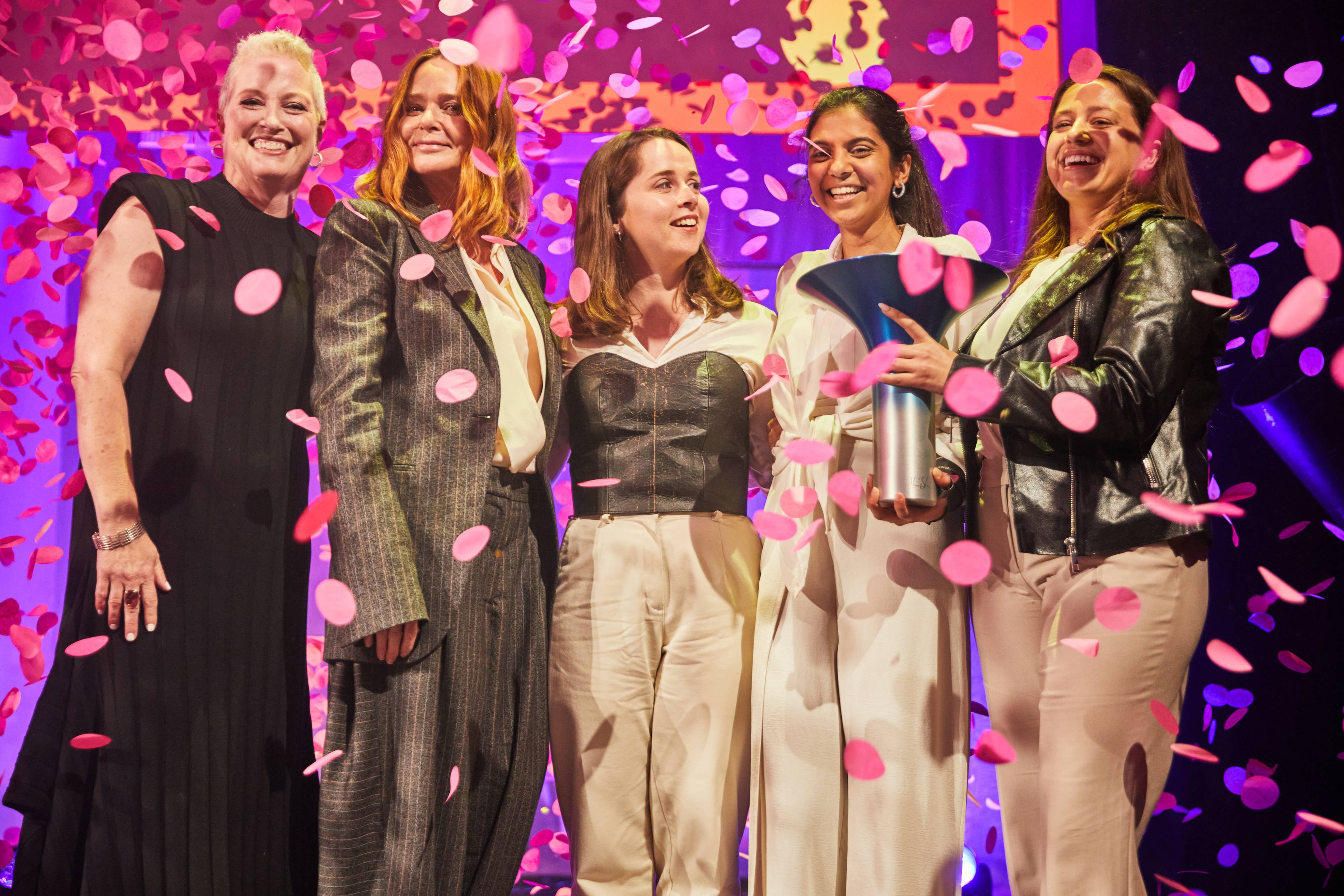 Five people celebrating on stage with pink confetti falling, holding an award. They are smiling and dressed in formal attire.