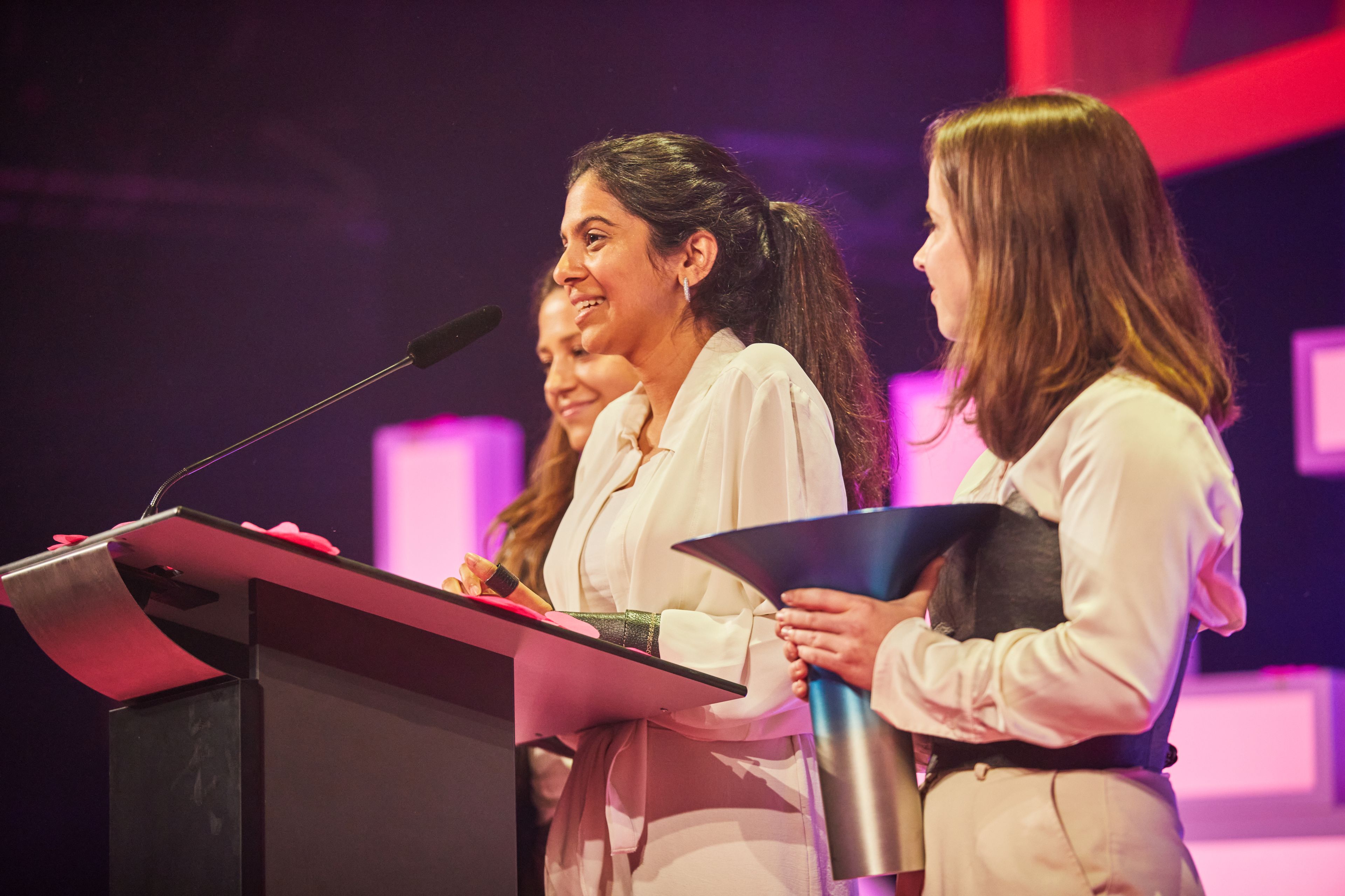 Three women stand on stage at a podium, one speaking into a microphone, with colorful lights in the background.