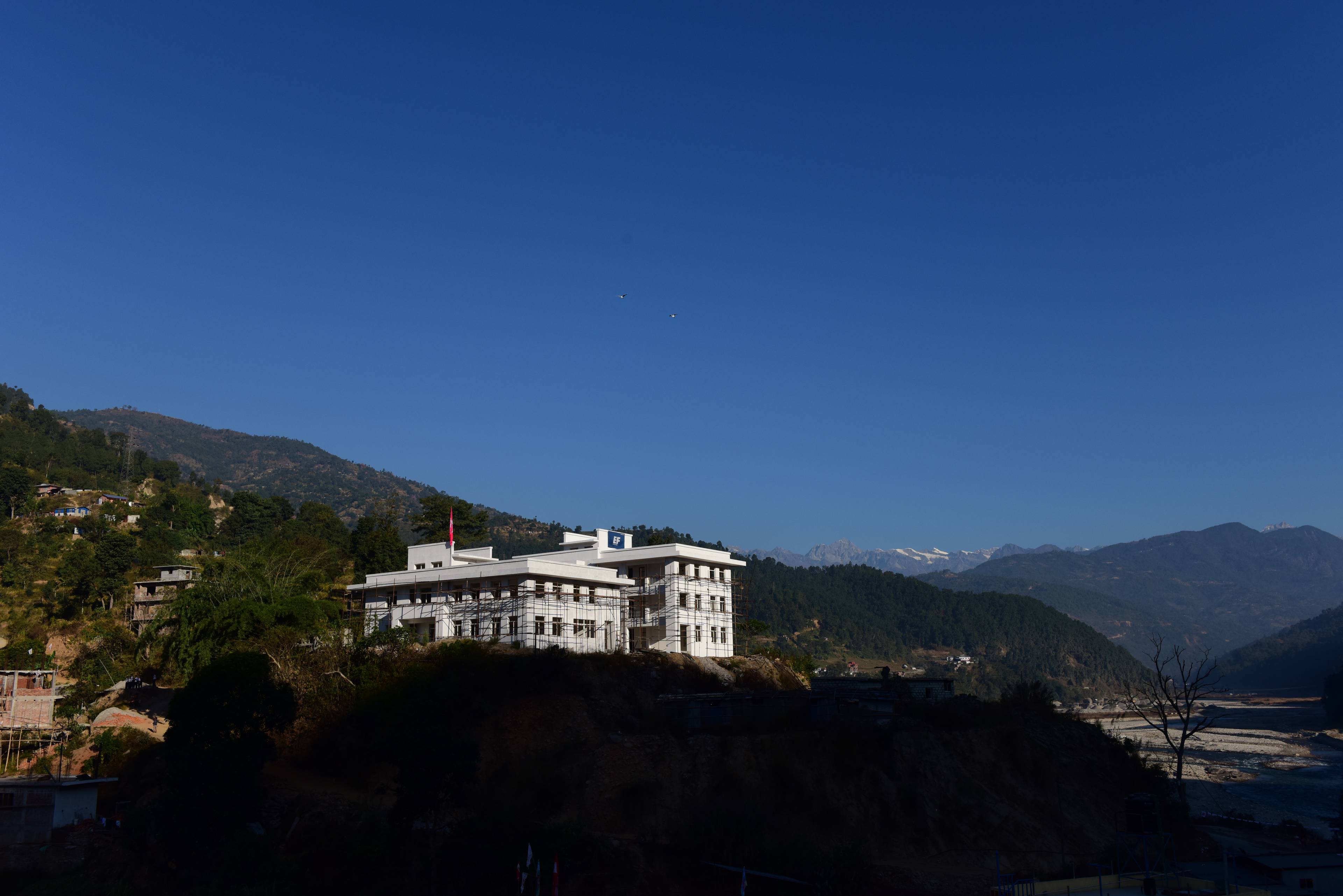 A white building on a hilltop surrounded by lush greenery and mountains under a clear blue sky.