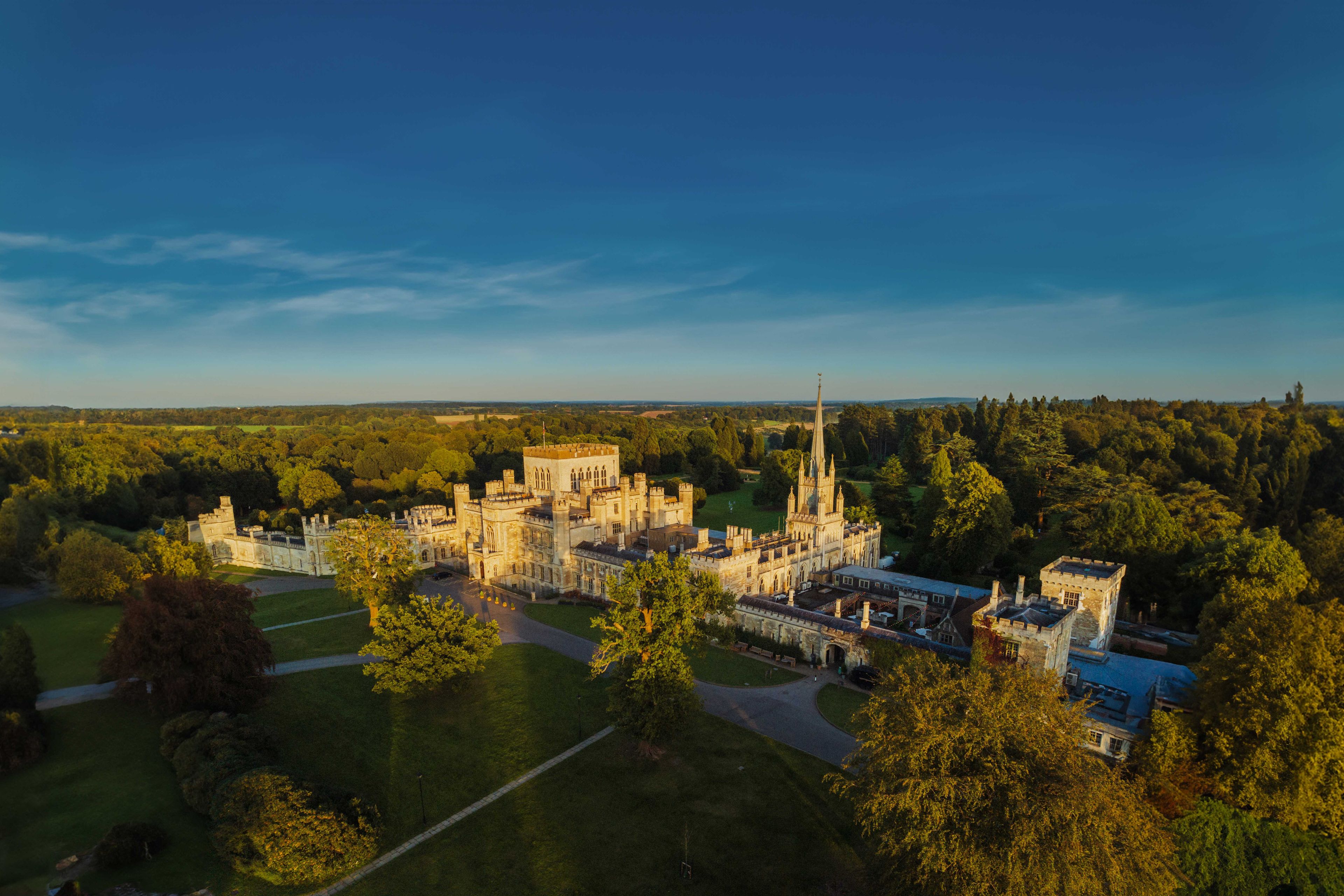 Aerial view of a large, historic castle surrounded by lush greenery and trees under a clear blue sky.