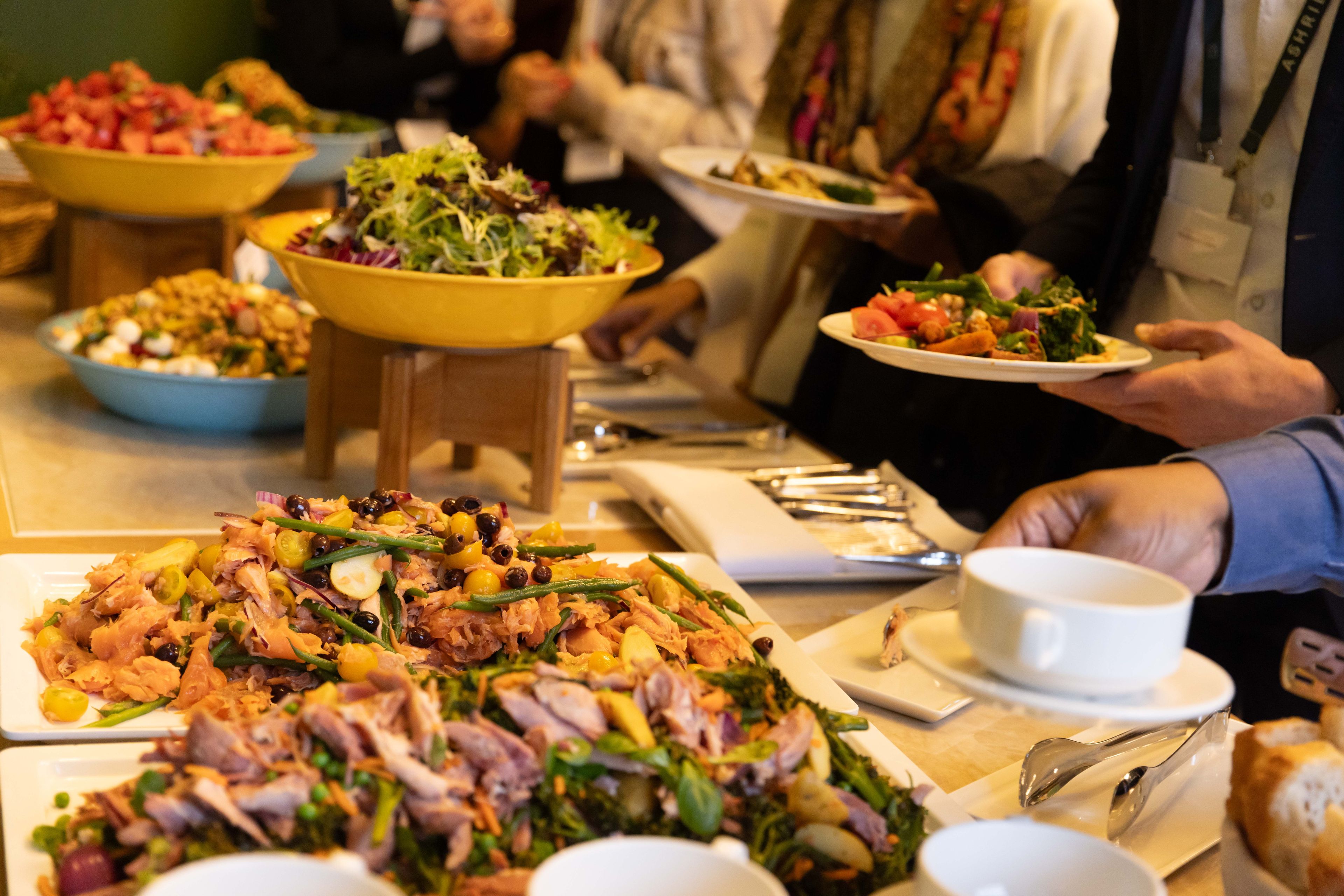 People serving themselves from a buffet with various salads and dishes, holding plates and cups, in a social gathering setting.