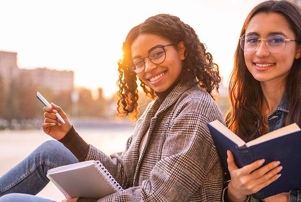 Two young women sitting outdoors, smiling, holding a notebook and a book, with a blurred cityscape in the background during sunset.