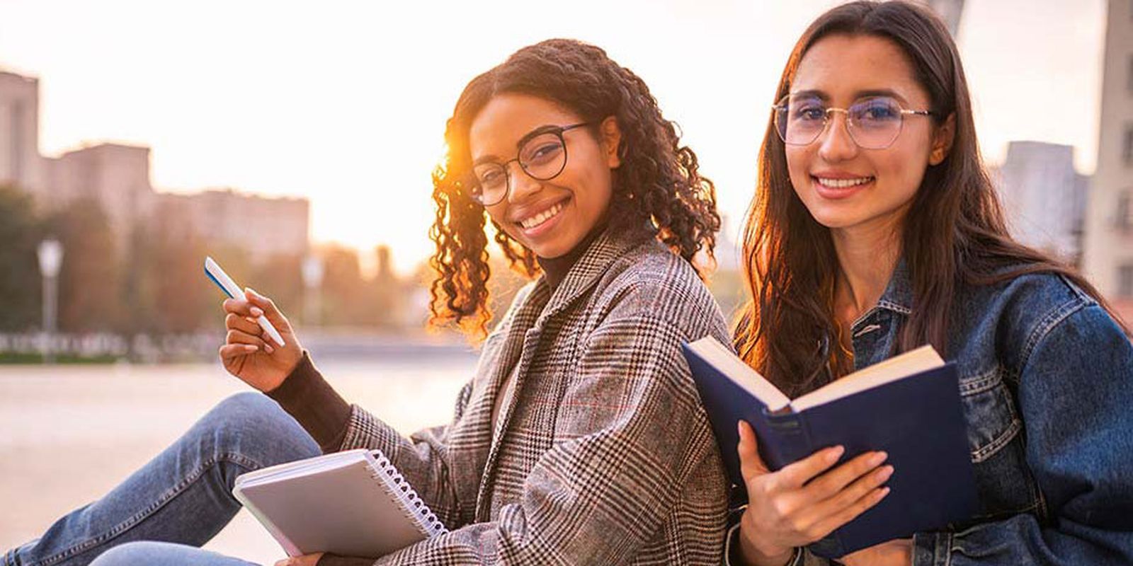 Two young women sitting outdoors, smiling, holding a notebook and a book, with a blurred cityscape in the background during sunset.