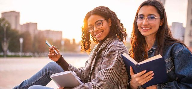 Two young women sitting outdoors, smiling, holding a notebook and a book, with a blurred cityscape in the background during sunset.