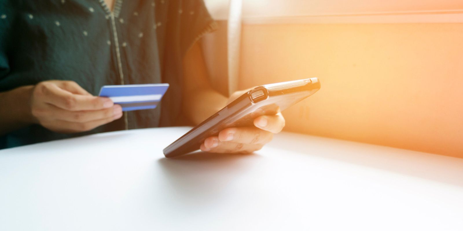 Person holding a smartphone and a credit card, sitting at a white table with soft sunlight in the background.