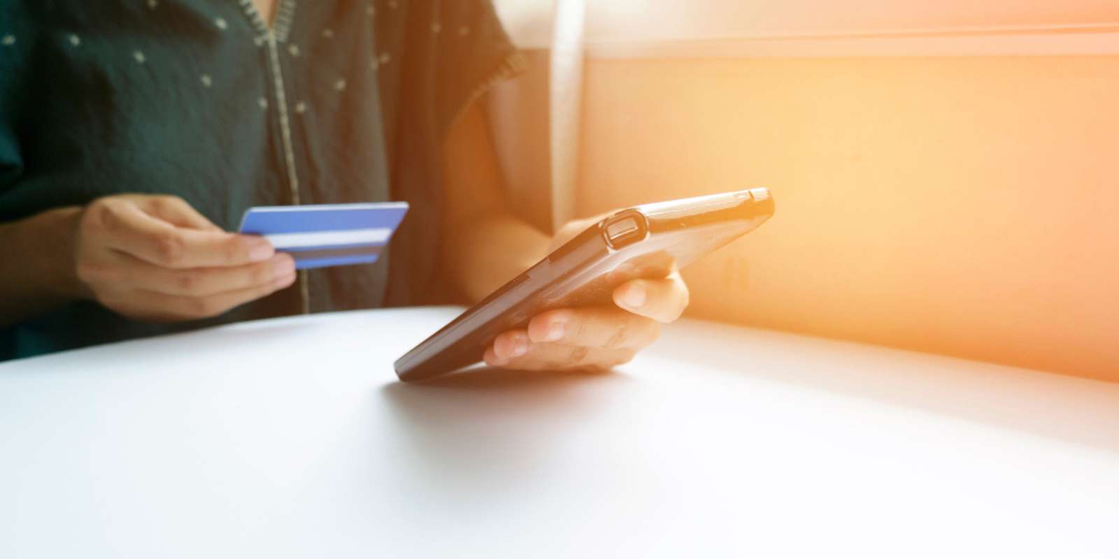 Person holding a smartphone and a credit card, sitting at a white table with soft sunlight in the background.