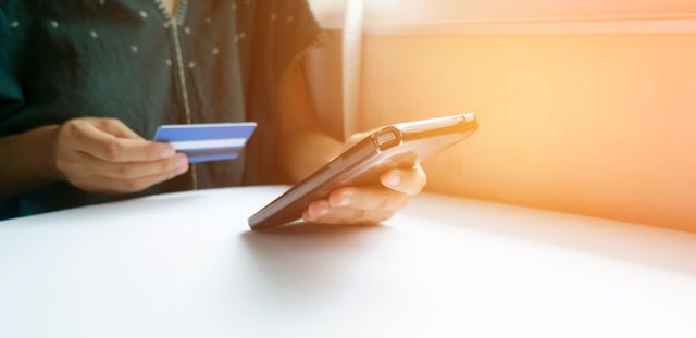 Person holding a smartphone and a credit card, sitting at a white table with soft sunlight in the background.