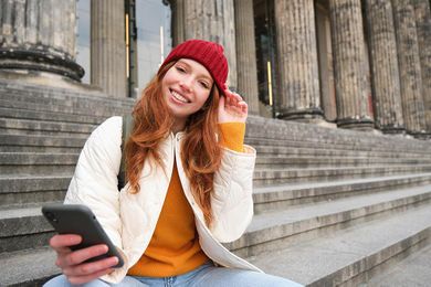 Woman in a red beanie and white jacket smiles while sitting on steps, holding a phone, with large stone columns in the background.