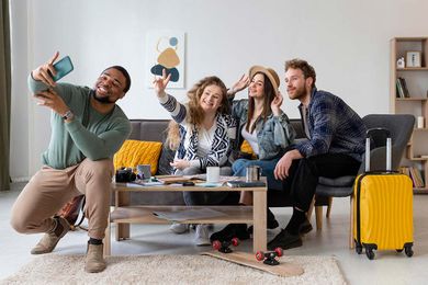 A group of four friends sitting in a living room, taking a selfie. The room has a couch, coffee table, and a yellow suitcase.