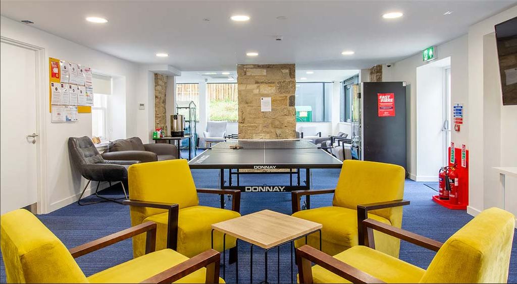 Recreation room with a ping pong table, yellow chairs around a small table, a sofa, and a coffee machine. Stone wall accent and blue carpet.