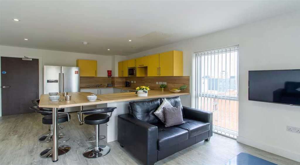 Modern kitchen and living area with yellow cabinets, a wooden island with bar stools, black sofa, wall-mounted TV, and large window with blinds.