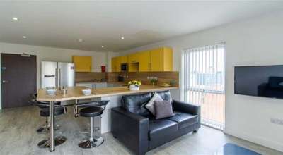 Modern kitchen and living area with yellow cabinets, a wooden island with bar stools, black sofa, wall-mounted TV, and large window with blinds.