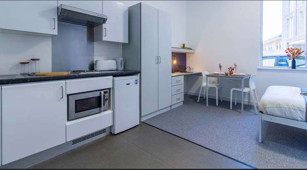 Modern studio apartment with a kitchenette, small dining area, bed, and a window. Features white cabinets, a microwave, and a mini fridge.