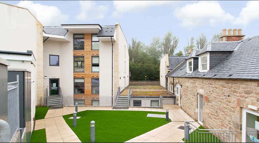 Modern building complex with a mix of stone and wood facade, surrounded by a courtyard with green artificial grass and paved walkways.