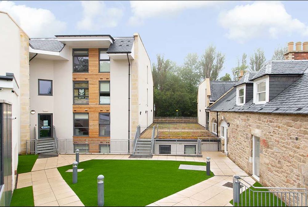 Modern building complex with a mix of stone and wood facade, surrounded by a courtyard with green artificial grass and paved walkways.