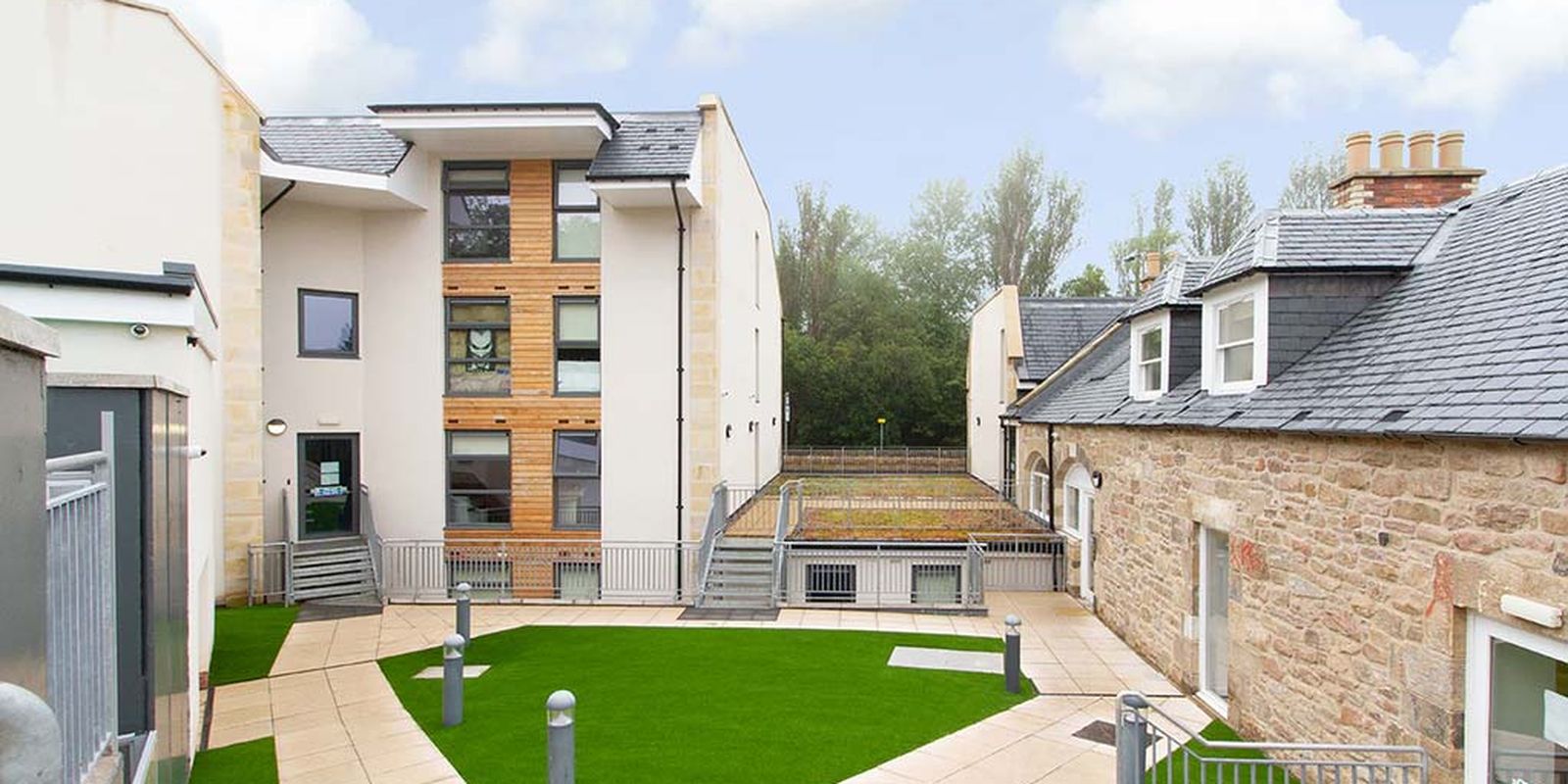 Modern building complex with a mix of stone and wood facade, surrounded by a courtyard with green artificial grass and paved walkways.