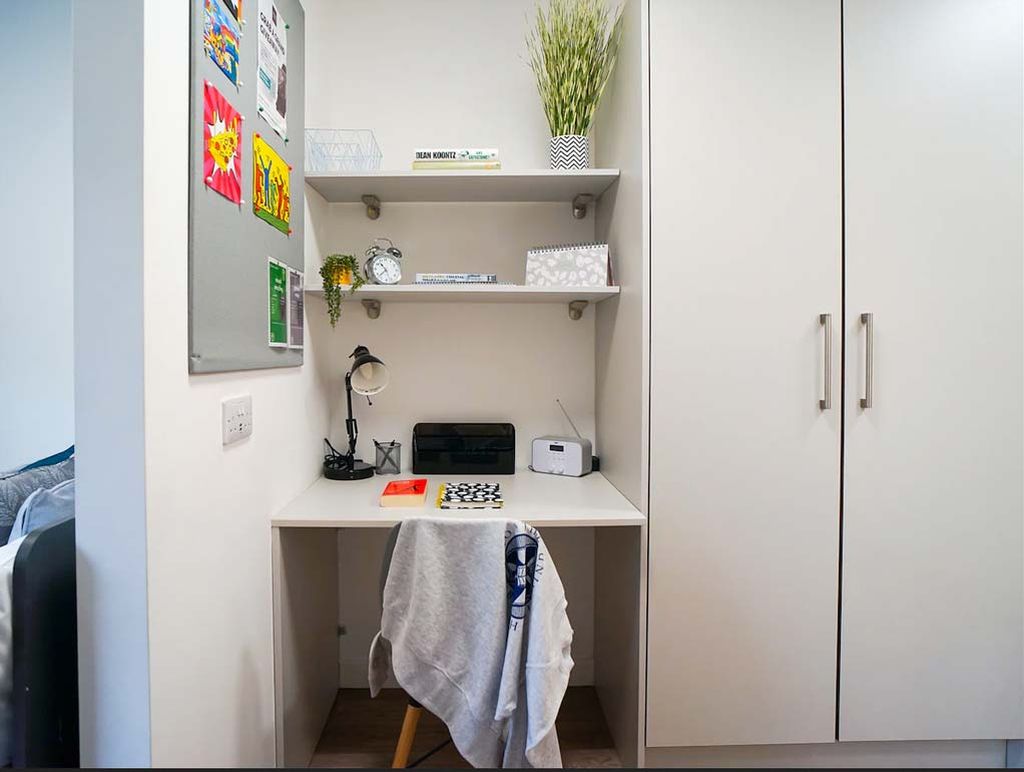 Compact study nook with a white desk, chair, shelves with decor, a bulletin board, and a large wardrobe. A towel drapes over the chair.