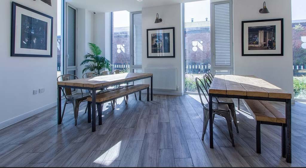Bright room with wooden tables, metal chairs, framed art on walls, large windows, and a potted plant. Natural light fills the space.