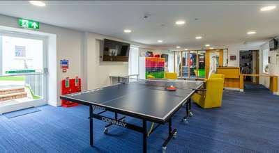 Room with a ping pong table, blue carpet, yellow chairs, TV, and colorful shelving. Exit signs and fire extinguishers are visible.