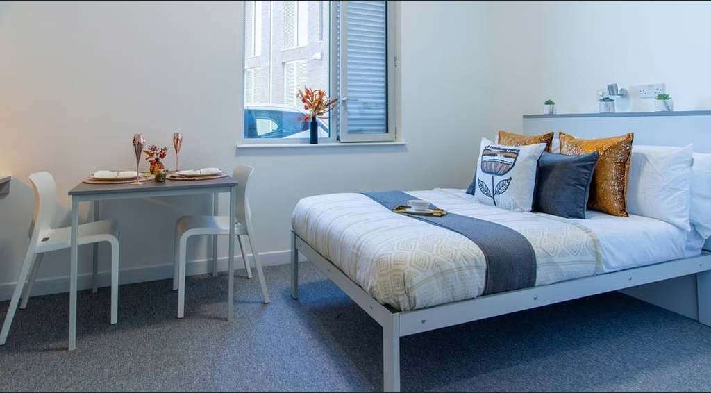Cozy bedroom with a neatly made bed, decorative pillows, a small dining table set for two, and a window letting in natural light.