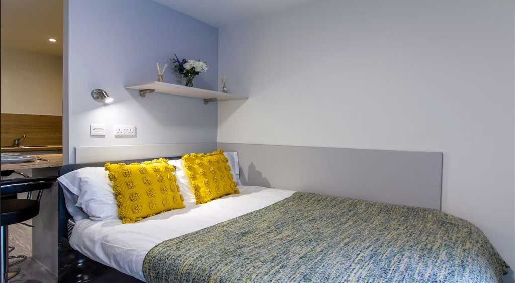 Cozy bedroom with a double bed, yellow pillows, a gray and green blanket, and a shelf with flowers above. A small kitchen area is visible to the left.