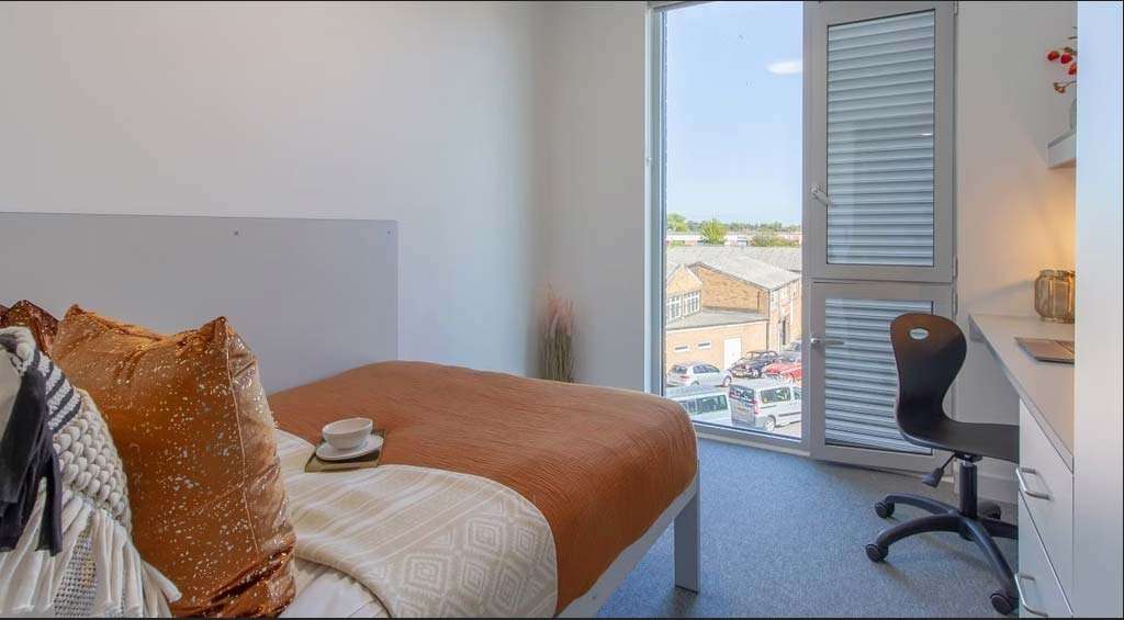 Modern bedroom with a neatly made bed, orange pillows, a desk with a chair, and a window overlooking a parking lot and buildings.
