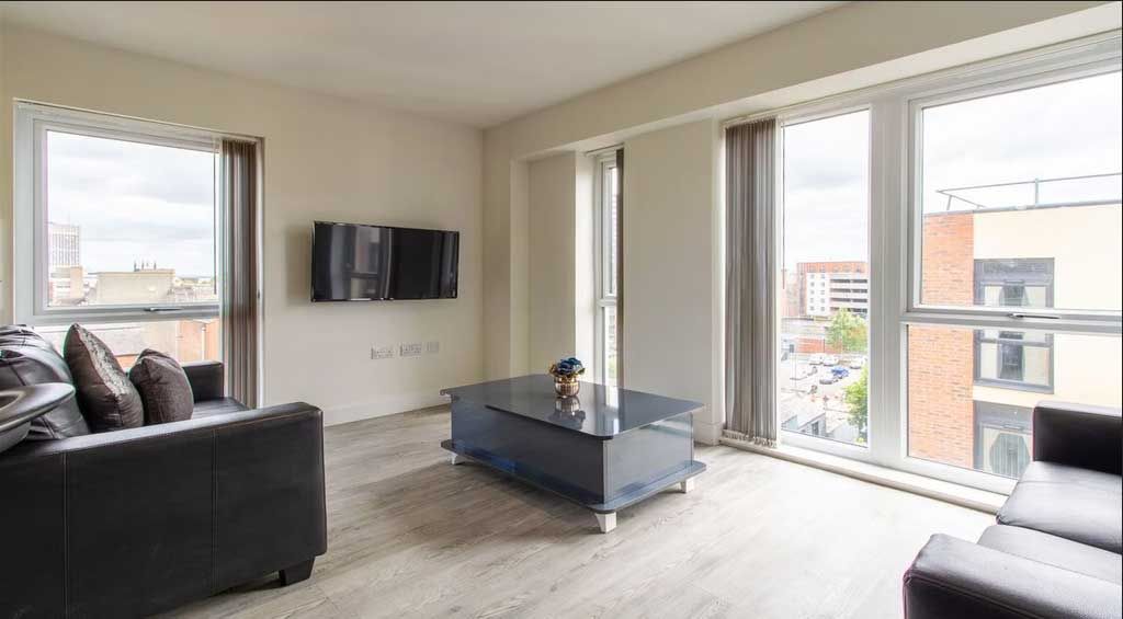 Modern living room with large windows, black sofa, wall-mounted TV, and a sleek glass coffee table on light wood flooring. Cityscape view outside.