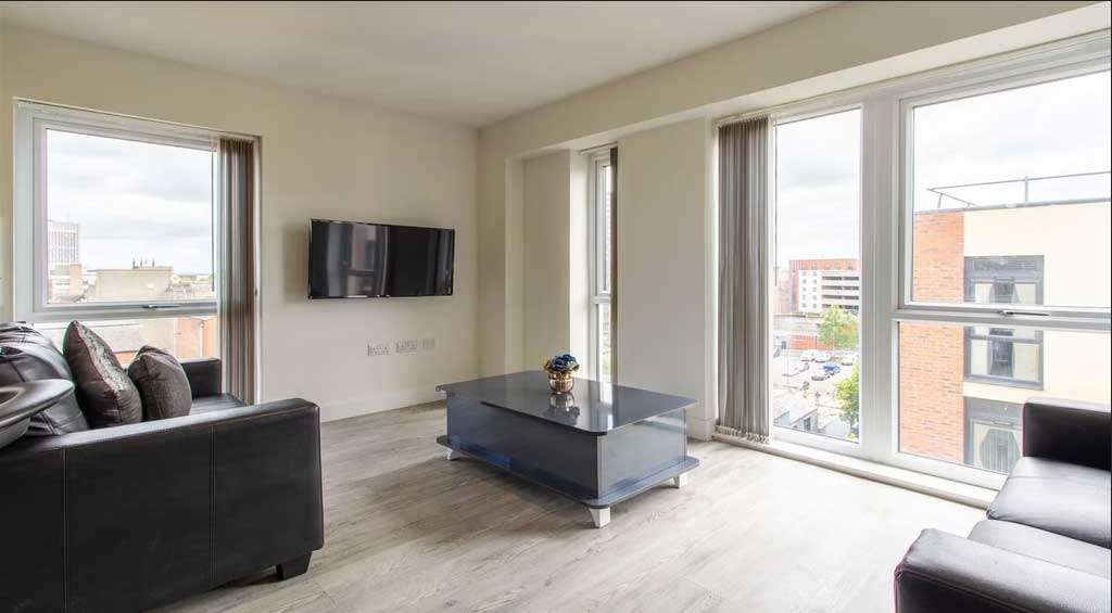 Modern living room with large windows, black sofa, wall-mounted TV, and a sleek glass coffee table on light wood flooring. Cityscape view outside.