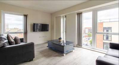 Modern living room with large windows, black sofa, wall-mounted TV, and a sleek glass coffee table on light wood flooring. Cityscape view outside.