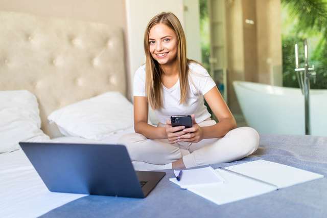 A smiling young woman sitting on a bed with a laptop, notepad, and smartphone, engaged in work or study in a bright, modern room.
