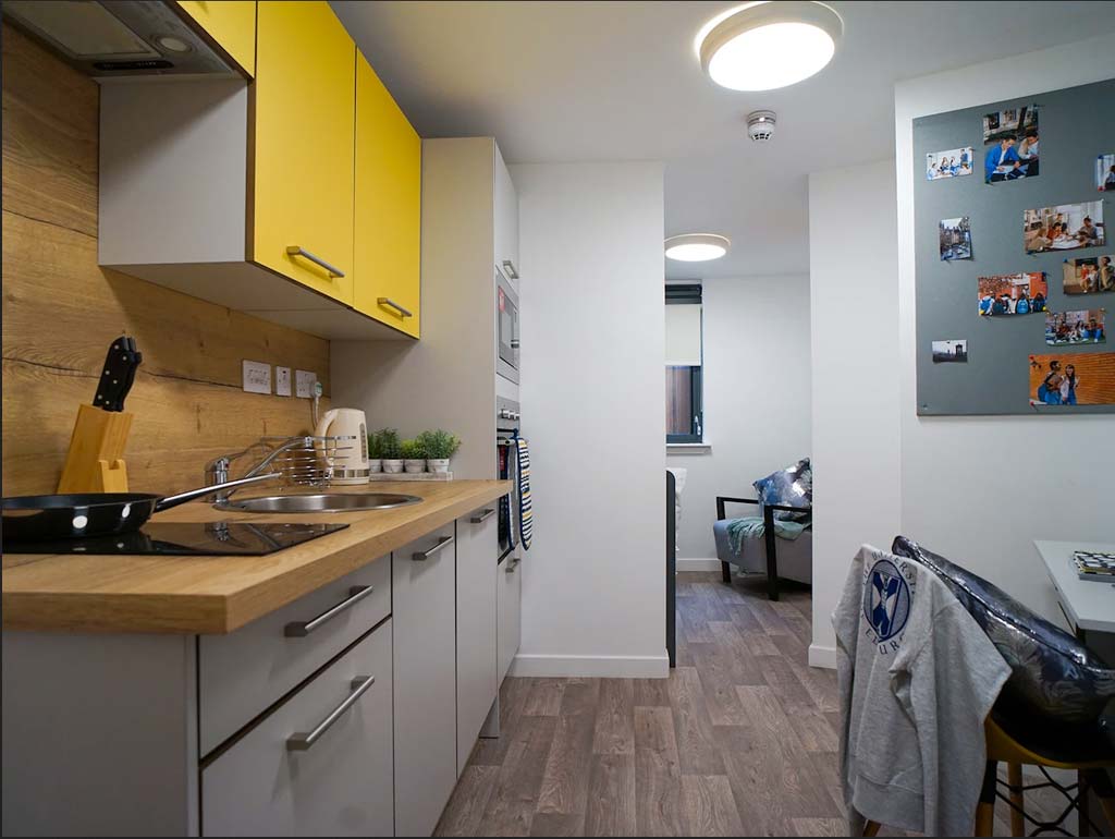 Modern kitchen with yellow cabinets, wooden countertop, and wall-mounted photos. A cozy living area is visible in the background.