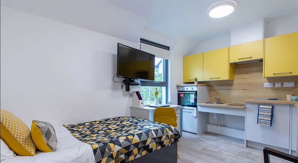 Modern studio apartment with a bed, geometric-patterned bedding, wall-mounted TV, yellow kitchen cabinets, and a small dining area by the window.