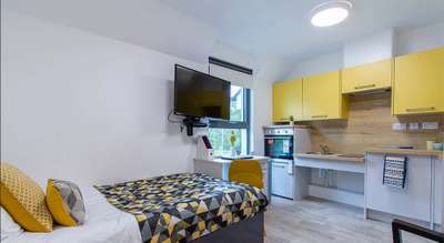 Modern studio apartment with a bed, geometric-patterned bedding, wall-mounted TV, yellow kitchen cabinets, and a small dining area by the window.
