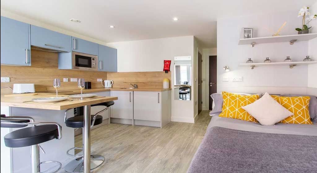 Modern studio apartment with a kitchenette, bar stools, and a bed with yellow and white pillows. Light wood flooring and bright lighting.