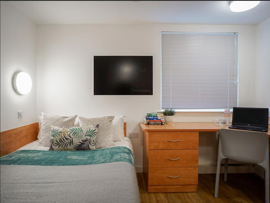A tidy bedroom with a bed, decorative pillows, a wall-mounted TV, a wooden desk with books, a laptop, and a chair under a window with blinds.