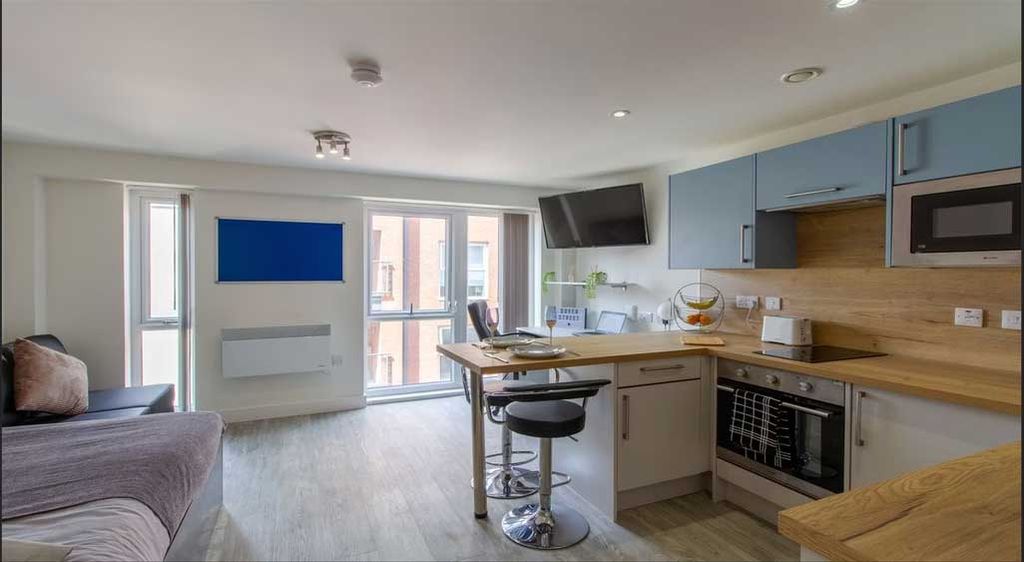 Modern studio apartment with a kitchenette, bar stool seating, wall-mounted TV, and large windows. Light wood flooring and neutral decor.