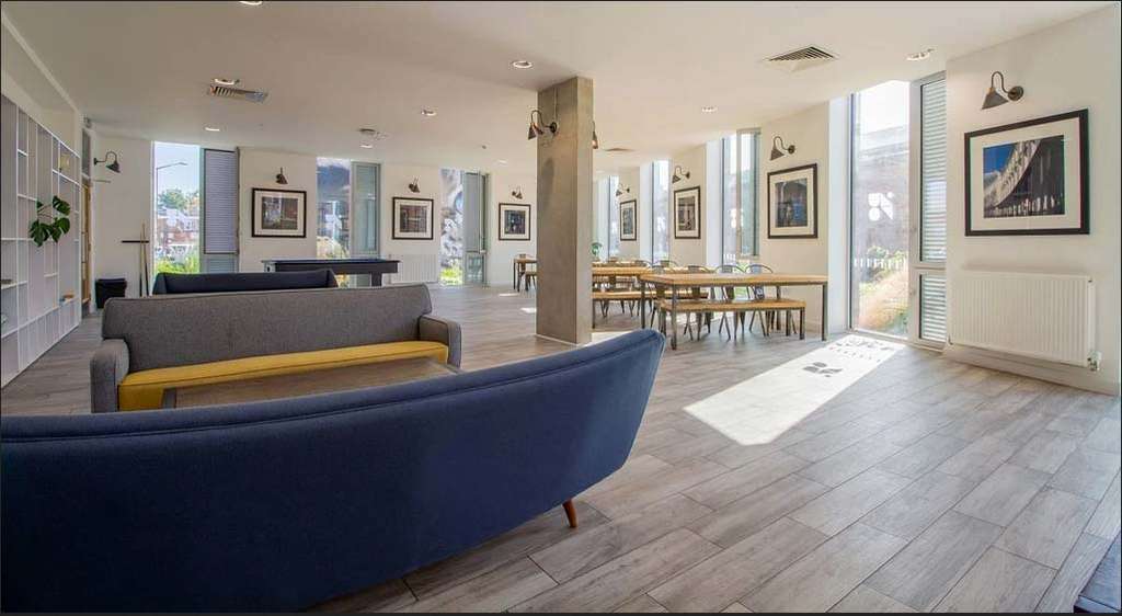 Modern lounge with blue sofas, wooden tables, and framed photos on the walls. Large windows allow natural light to fill the room.