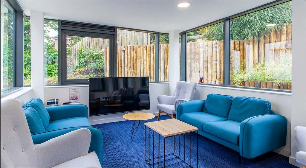 Modern living room with large windows, blue sofas, a flat-screen TV, and a wooden coffee table. Outside, a wooden fence and greenery are visible.