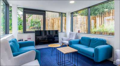 Modern living room with large windows, blue sofas, a flat-screen TV, and a wooden coffee table. Outside, a wooden fence and greenery are visible.