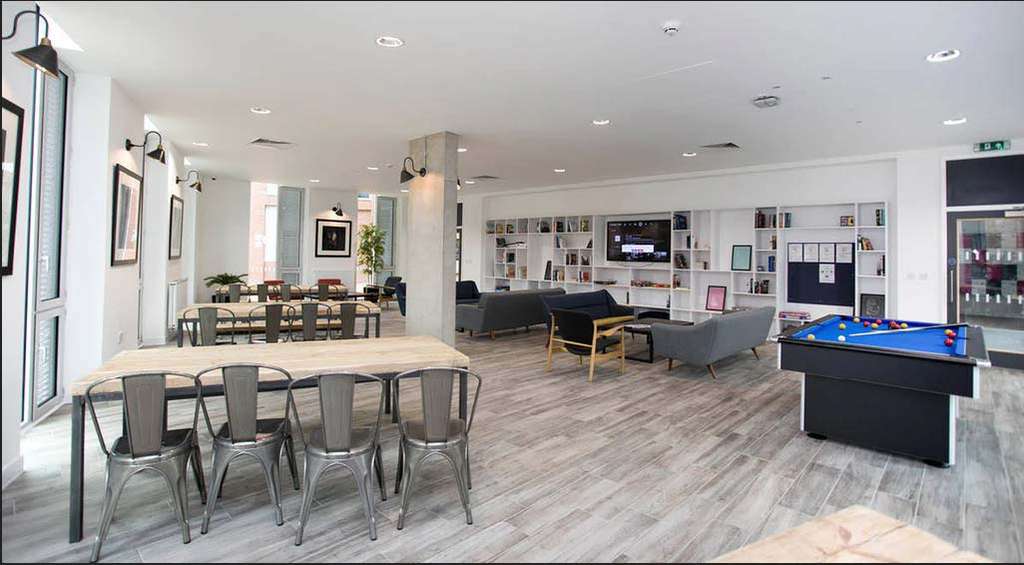 Modern communal lounge with a pool table, sofas, bookshelves, and a dining area with metal chairs on a wood-patterned floor.