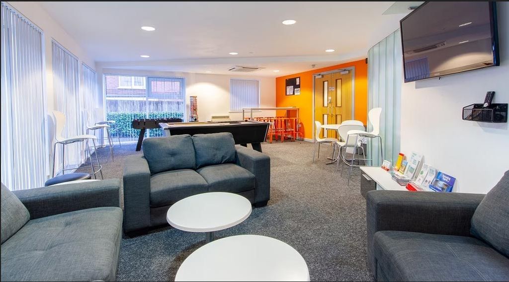 Modern lounge with gray sofas, round white tables, a foosball table, bar stools, and orange accent wall. Bright windows and brochures on display.