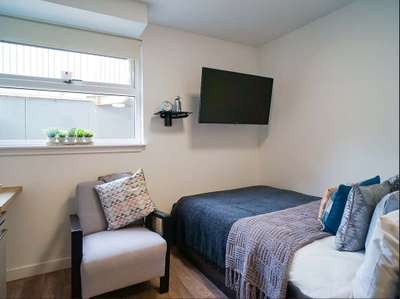 Cozy room with a bed, wall-mounted TV, armchair, and window with potted plants. Neutral tones and a simple, modern design.
