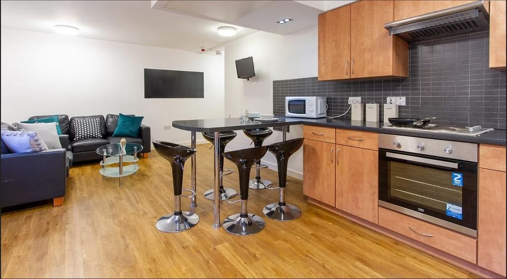 Modern kitchen and living area with wooden flooring, black bar stools, sleek black backsplash, and a small seating area with a sofa and glass table.