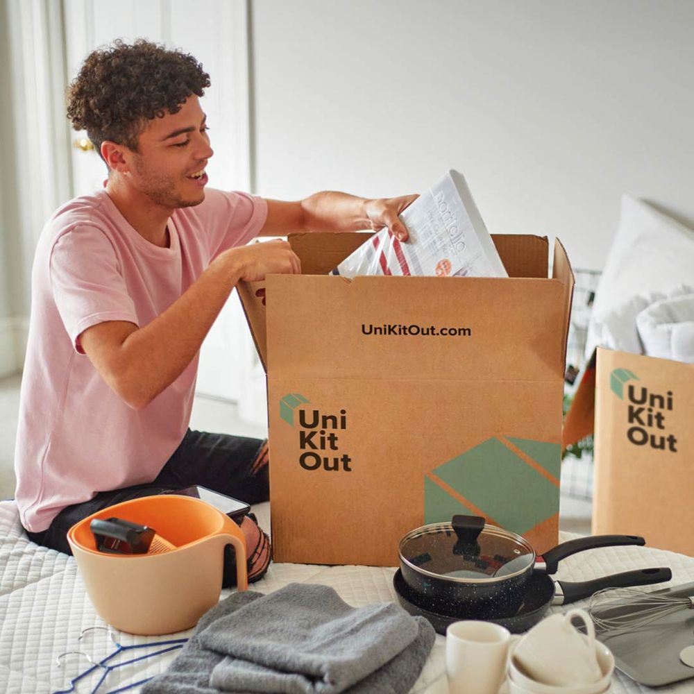 A smiling person opens a Uni Kit Out box, surrounded by household items like a bowl, towel, and pan, suggesting a move or unpacking at home.