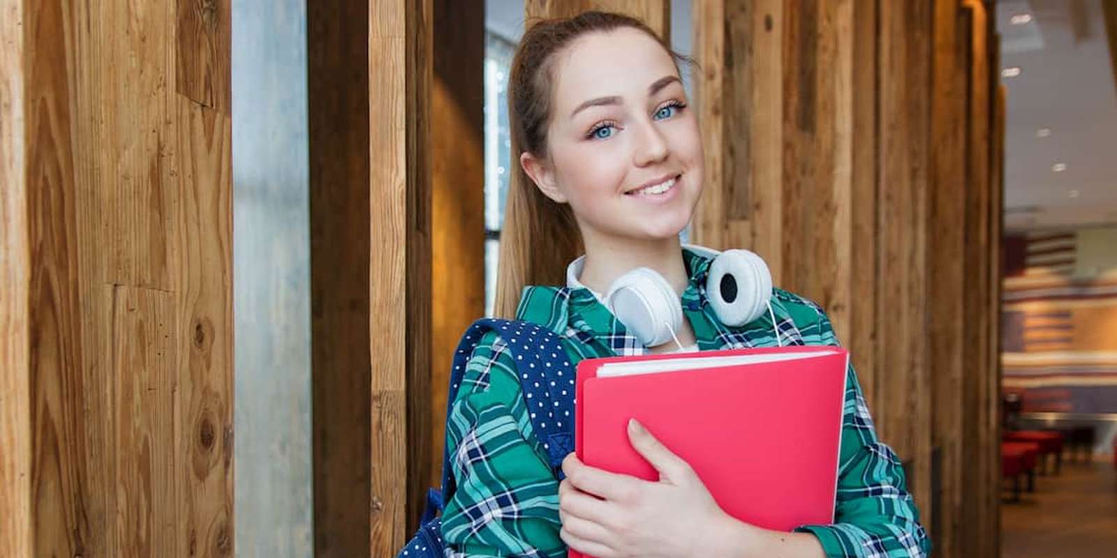 A young student with a backpack and headphones around her neck, smiling and holding a red folder, stands in a modern wooden-paneled hallway.