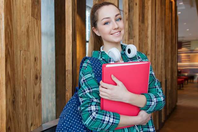 A young student with a backpack and headphones around her neck, smiling and holding a red folder, stands in a modern wooden-paneled hallway.