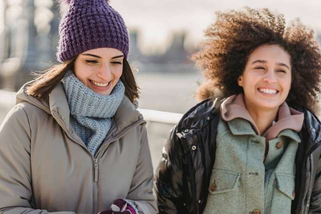 Two women happily walking outdoors on a sunny, chilly day, one wearing a purple beanie and scarf, the other with a curly hair, both smiling and enjoying each other's company.
