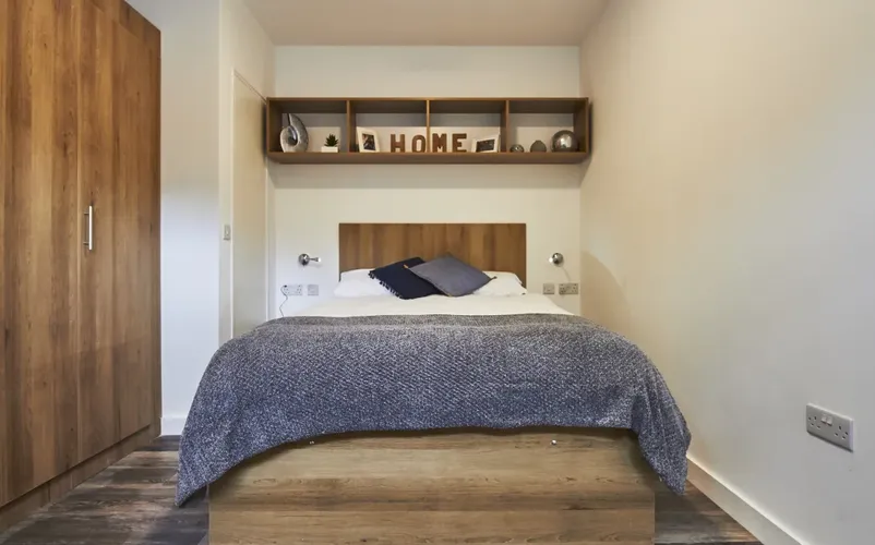 Cozy bedroom with a wooden bed, gray blanket, and decorative shelf above featuring letters spelling "HOME." Wardrobe on the left.