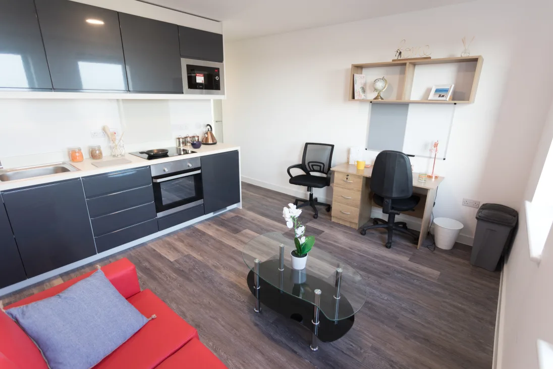 Modern studio apartment with a kitchenette, black cabinets, red sofa, glass coffee table, and two desks with chairs on wooden flooring.